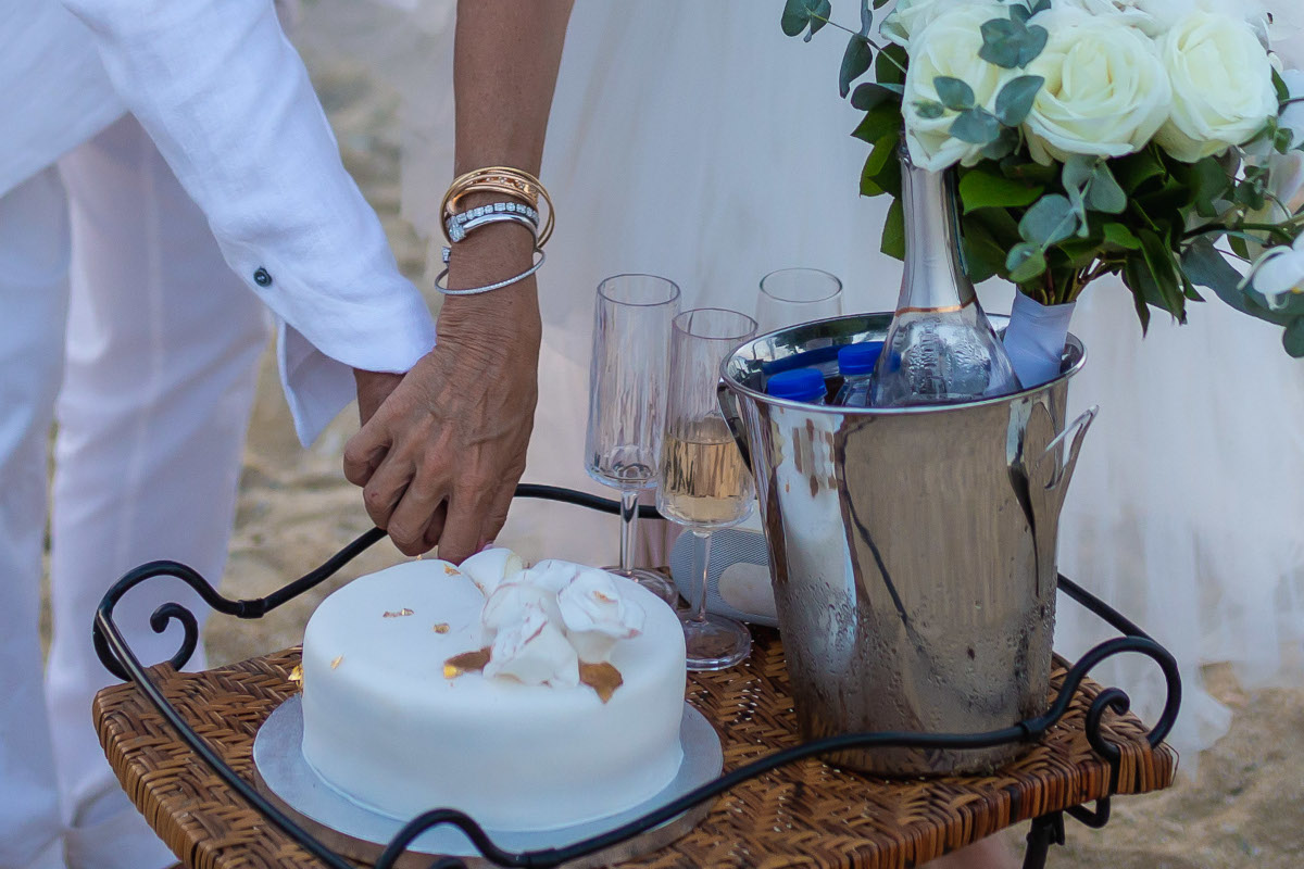 A couple reaches for their wedding cake on a tray, surrounded by champagne flutes and a bouquet of white roses on their Mykonos beach wedding.