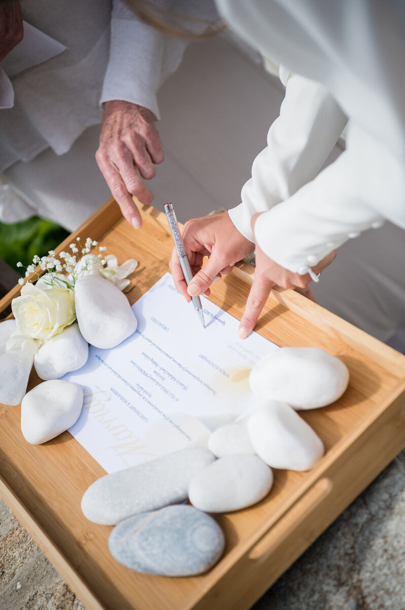 A close-up of hands signing a document on a wooden tray adorned with white stones and a white rose, capturing a meaningful moment of their Mykonos wedding.