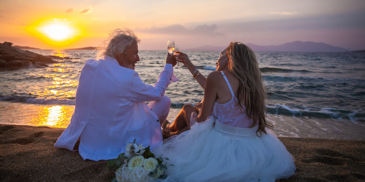 Newlywed couple sitting on the beach enjoying Mykonos sunset with bubly, in this mykonos wedding package