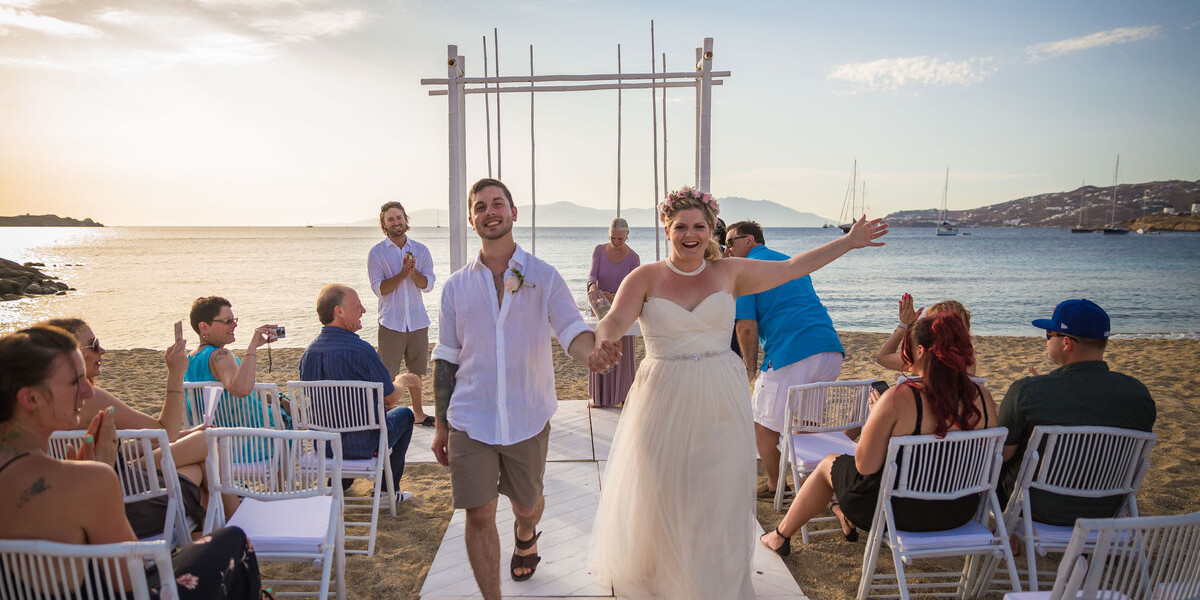 A bride and groom joyfully walk down a sandy beach aisle, celebrating their love in a picturesque Mykonos ceremony.
